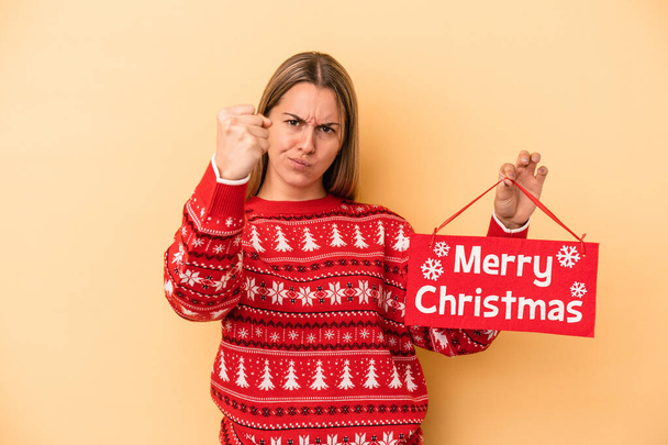 Young caucasian woman holding a merry christmas placard isolated on yellow background showing fist to camera, aggressive facial expression. - Foto, immagini