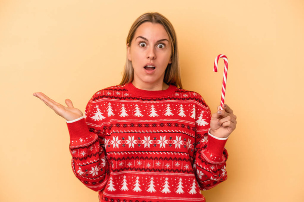 Mladá běloška žena drží vánoční hůl izolované na žlutém pozadí překvapené a šokované. - Fotografie, Obrázek