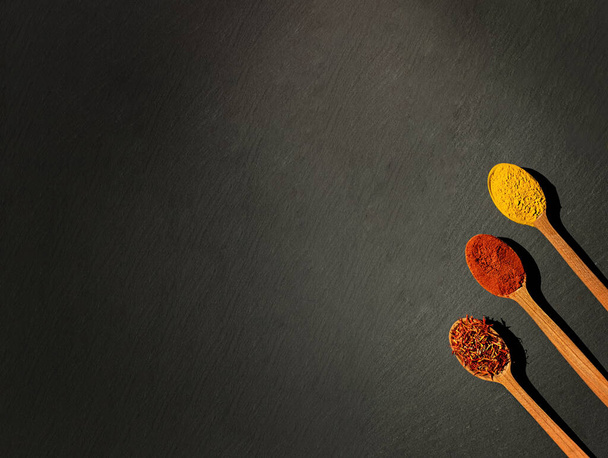 On a black background, three wooden spoons with spices, bell pepper, chili and curry, turmeric. - Valokuva, kuva