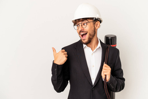 Young mixed race architect man wearing a helmet isolated on grey background points with thumb finger away, laughing and carefree. - Foto, Imagem