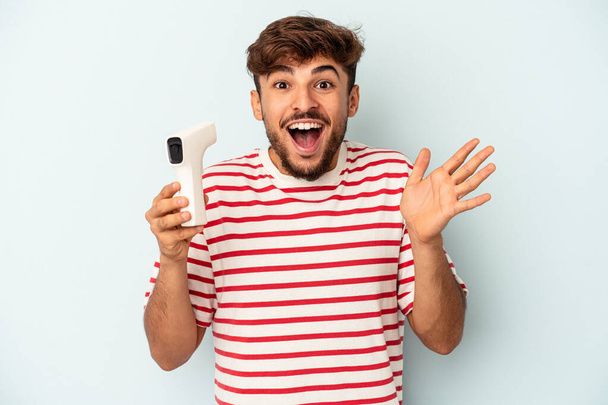 Young mixed race man holding thermometer isolated on blue background receiving a pleasant surprise, excited and raising hands. - Foto, Imagem