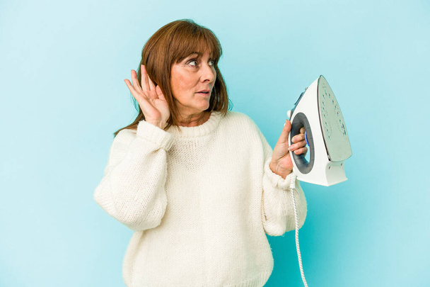 Middle age caucasian woman holding a iron isolated on blue background trying to listening a gossip. - Фото, изображение