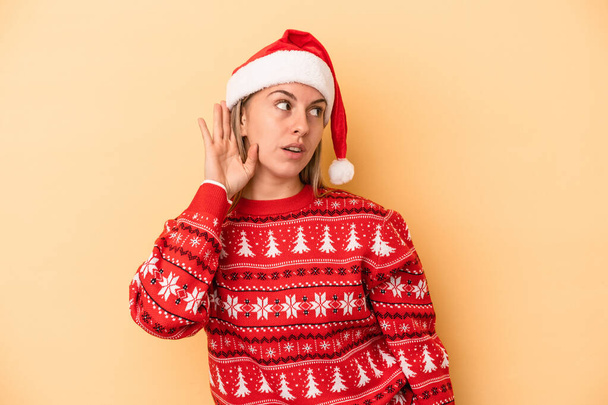 Νεαρή λευκή γυναίκα γιορτάζει τα Χριστούγεννα απομονωμένη σε κίτρινο φόντο προσπαθώντας να ακούσει ένα κουτσομπολιό. - Φωτογραφία, εικόνα