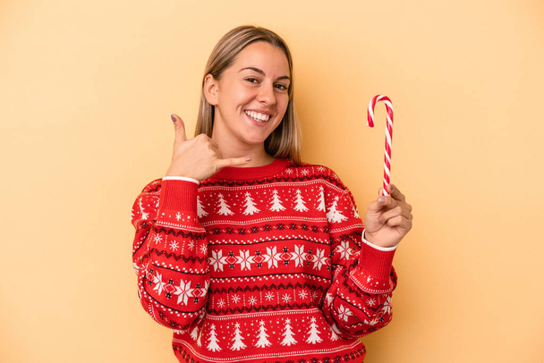 Young caucasian woman holding a christmas stick isolated on yellow background showing a mobile phone call gesture with fingers. - Photo, Image