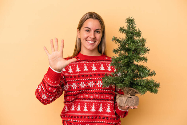 Молода біла жінка тримає маленьку різдвяну ялинку ізольовано на жовтому тлі усміхнений веселий показ п'ятої номери з пальцями
. - Фото, зображення