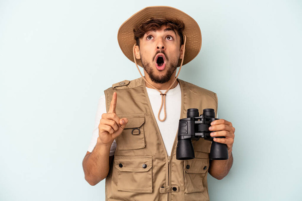 Young mixed race man holding binoculars isolated on blue background pointing upside with opened mouth. - Фото, изображение