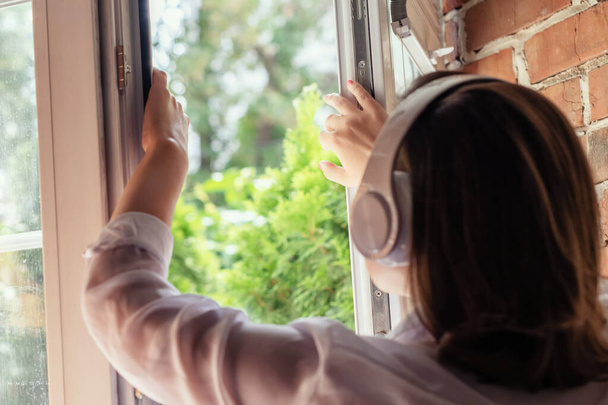 Young beautiful happy woman in headphones listening music opens  window of country house and breathes fresh air in the early summer morning. Back view. - Foto, Imagen