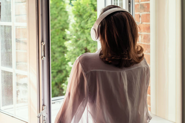 Young beautiful woman in headphones listening music opens of the bathroom window of country house and breathes fresh air in the early summer morning. Back view. - Foto, immagini