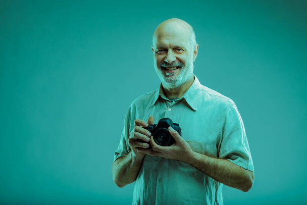 Confident friendly senior photographer smiling at the camera as he holds his camera in his hands over a blue-grey studio background - Фото, зображення