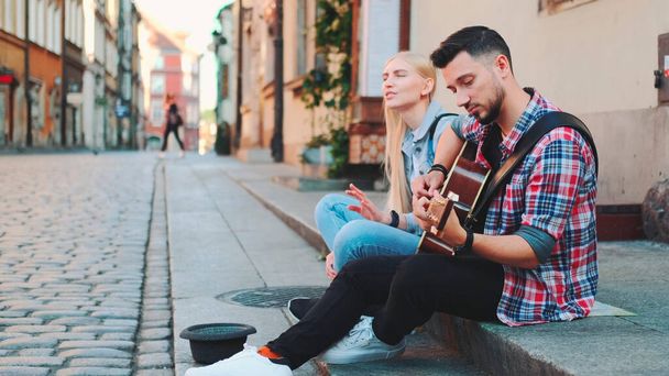 Νεαροί τραγουδιστές κάθονται στο πεζοδρόμιο, παίζουν κιθάρα και τραγουδούν - Φωτογραφία, εικόνα