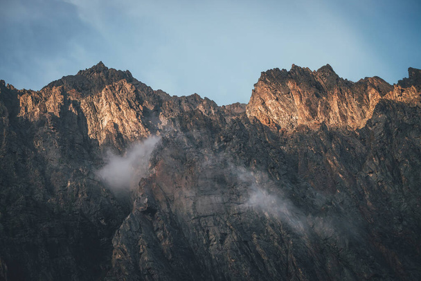 Ορεινό τοπίο με βράχια στο χρυσό φως του ήλιου και χαμηλά σύννεφα. Φοβερός βραχώδης τοίχος με αιχμηρή κορυφή σε χρυσό ήλιο σε συννεφιασμένο καιρό. Ατμοσφαιρικό τοπίο με ψηλό βραχώδες βουνό κορυφή στα σύννεφα - Φωτογραφία, εικόνα