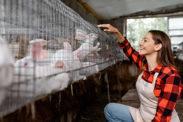 Young happy woman farmer is looking at the metal cages with white rabbits in her farm. The farmer takes care of the animals. Bio meat concept  - Fotografie, Obrázek