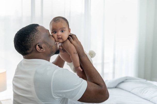 African father holding and kissing daughter's cheek, is baby newborn 3 month, in white bed room, to relationship of African familiy and baby newborn concept. - Fotografie, Obrázek