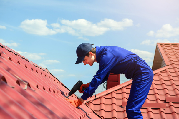 Close-up van de jonge happy man aannemer in blauwe overall is het repareren van een rood dak met elektrische schroevendraaier. Hij glimlacht. Dakbedekking concept. - Foto, afbeelding