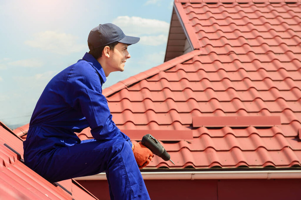 Young happy man contractor worker in blue overalls is repairing a red roof with electric screw driver and have a rest on the roof. Roofing concept. Copy space for text - Foto, imagen