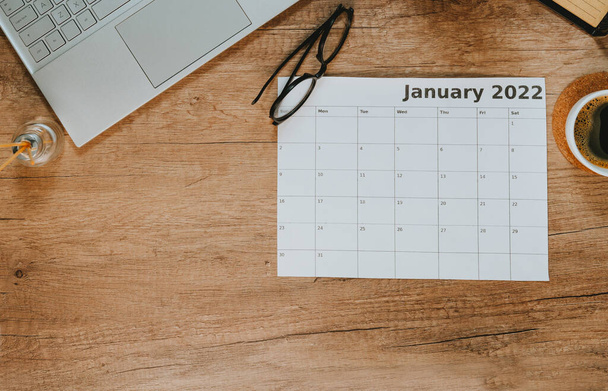 Top view of laptop and calendar of January 2022 on wooden background with blank space for text. Flat lay of workspace. Planning concept. - 写真・画像