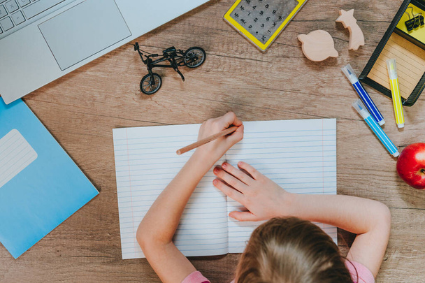 Dívka sedící u stolu a cvičící v zápisníku. Pohled shora, plochý. Dřevěný stůl s notebookem, notebookem, kalkulačkou a šablonou. Koncept vzdělávání. - Fotografie, Obrázek