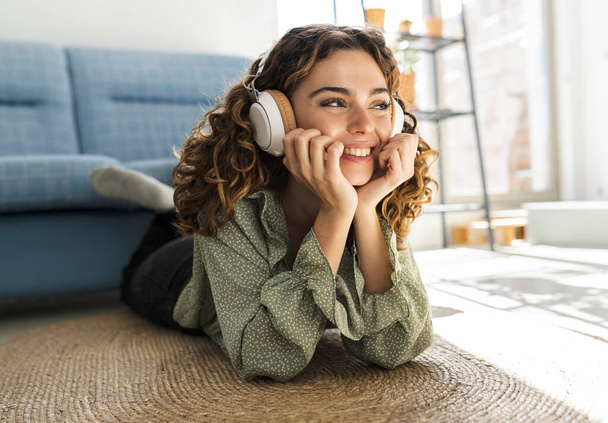 pretty curly-haired woman lying on the floor at home listening to music happy smiling relaxed - Photo, Image