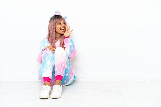 Young mixed race woman wearing a unicorn pajama sitting on the floor isolated on white background shouting with mouth wide open to the lateral - Foto, immagini