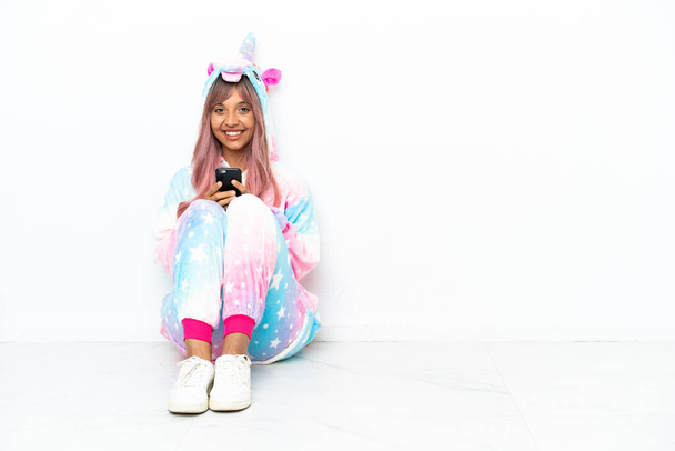 Young mixed race woman wearing a unicorn pajama sitting on the floor isolated on white background sending a message with the mobile - Foto, Imagem