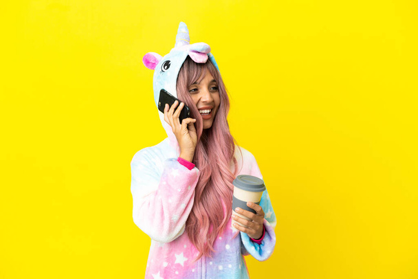 Joven mujer de raza mixta con un pijama unicornio aislado sobre fondo blanco sosteniendo café para llevar y un móvil - Foto, Imagen