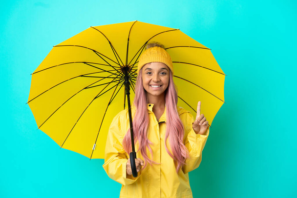 Young mixed race woman with rainproof coat and umbrella showing and lifting a finger in sign of the best - Valokuva, kuva