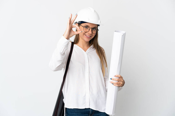 Young Lithuanian architect woman with helmet and holding blueprints isolated on white background showing ok sign with fingers - Fotó, kép