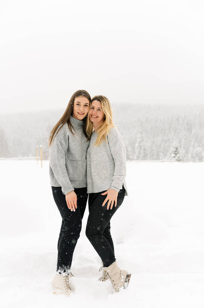 Mother and dauther in grey sweathers walks in a snowfall. Woman and girl cuddling in the winter forest. Outdoor portrait of beauty women posing in wood. family concept - Fotoğraf, Görsel