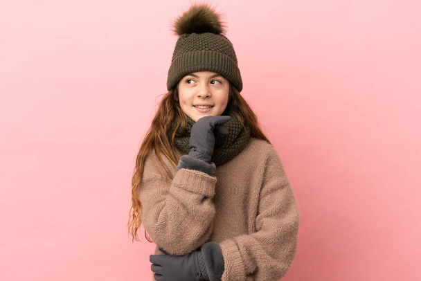 Kış şapkalı küçük kız pembe arka planda izole edilmiş ve yukarı bakıyor. - Fotoğraf, Görsel