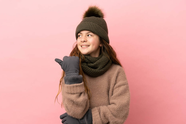Menina com chapéu de inverno isolado no fundo rosa apontando para o lado para apresentar um produto - Foto, Imagem
