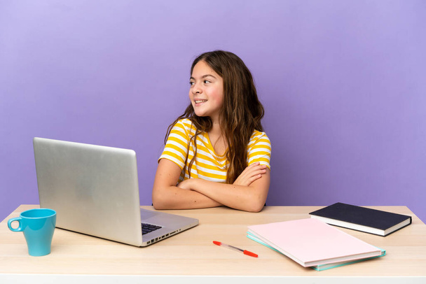 Little student girl in a workplace with a laptop isolated on purple background with arms crossed and happy - Fotó, kép