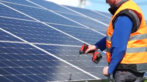 Solar power plant worker standing on a stepladder tightens solar panels fixing bolts with a drill - Materiał filmowy, wideo