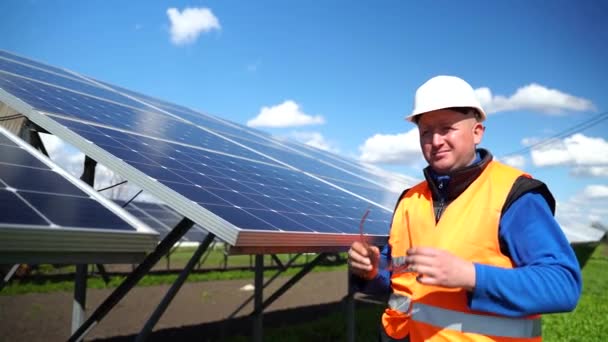 Portrait of engineer in uniform who putting on protective goggles while standing against the background of solar panels at a power station - Filmati, video