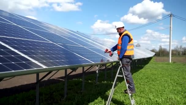 Solar power plant worker standing on a stepladder tightens solar panels fixing bolts with a drill - Video