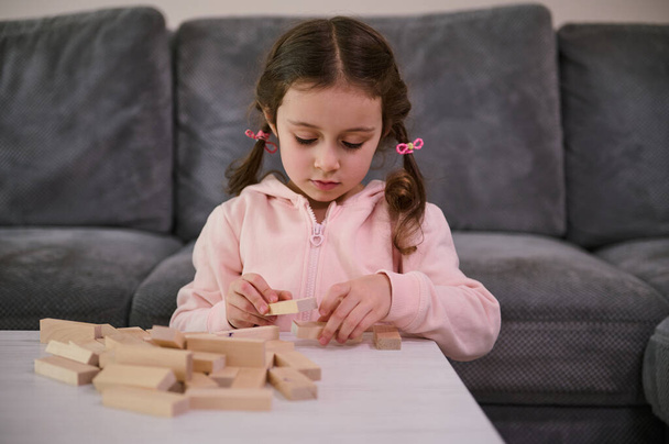 Education, development of fine motor skills, developmental board games concept. Adorable European girl concentrated on building with wooden blocks, sitting at a table in the living room - Zdjęcie, obraz