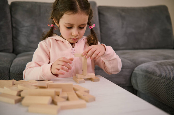 Adorable little girl with two pigtails, dressed in pink sweatshirt playing board game, building wooden constructions with blocks. Hand movement control and building computational skills concept. - Zdjęcie, obraz