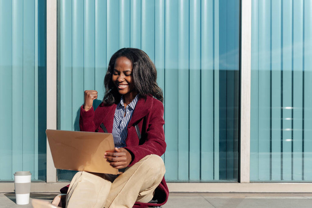 молода африканська студентка святкує успіх своїх університетських іспитів. Чорна дівчина використовує ноутбук, що сидить на вулиці
. - Фото, зображення