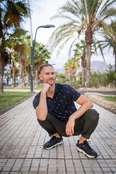 Guy squats outdoor in the middle of the sidewalk, he is smiling, happy and carefree. Portrait of a 30 year old bisexual caucasian man with copy space above him - Zdjęcie, obraz
