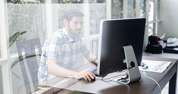 Bilgisayarla evden çalışan yakışıklı beyaz adamın camından bak. Ev konseptinde çalış. Genç adam evdeki şirket için çalışıyor.. - Fotoğraf, Görsel