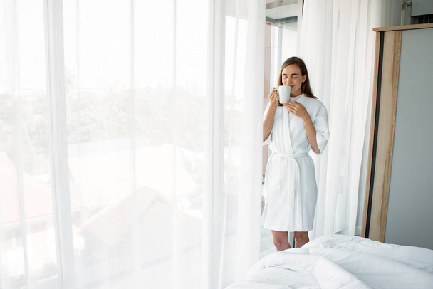 Красивая молодая кавказка в пижаме с бокалом белого кофе, готовая выпить у окна в спальне после утреннего пробуждения. Женщина, стоящая и пьющая кофе у окна
. - Фото, изображение