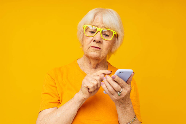 elderly woman in casual t-shirt communication by phone yellow background - Foto, Bild