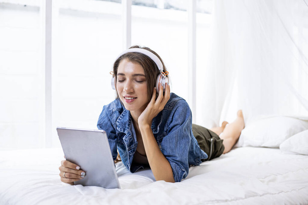 Молодая кавказская женщина в наслаждаясь онлайн музыкой на планшете во время отдыха на кровати с счастливым у себя дома в спальне. - Фото, изображение