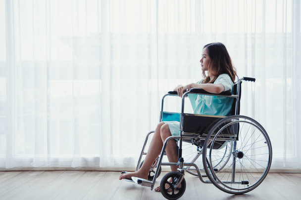 Woman patient in green shirt sitting on hospital clinic wheelchair in patient's room. Young female patient is recuperating in a hospital room due to illness. - 写真・画像