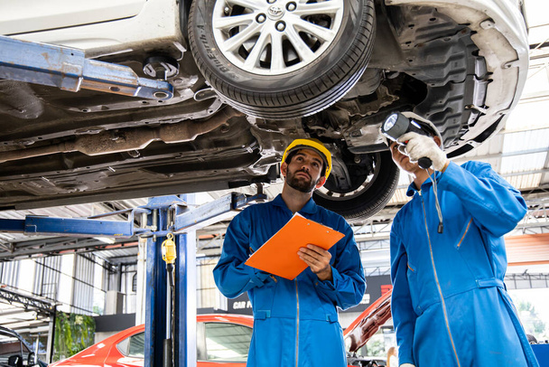 Two mechanic was checking the car suspension. Vehicle raised on lift at maintenance station. Car mechanic working at automotive service center. - 写真・画像