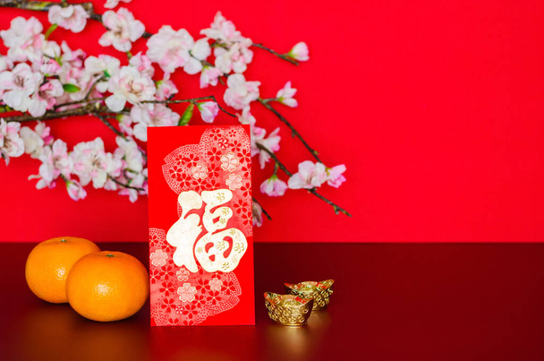 Red envelope packet or ang bao (word mean wealth) puts with oranges and ingots with peach blossom on red glitter paper background.  - Foto, afbeelding
