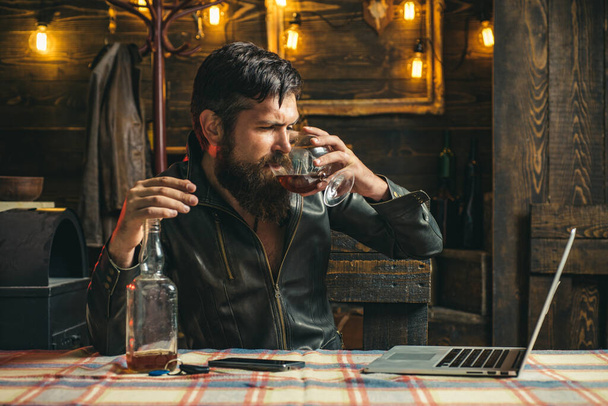 Drunk bearde sad pensive business man drinking whiskey in front of laptop at loft work place. Man or businessman drinks wiskey at home. Wasted alcoholic businessman drinking whiskey working at laptop. - Foto, Imagen