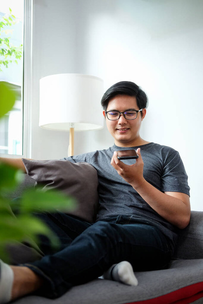 Un jeune homme souriant assis dans le salon et parlant sur un téléphone intelligent. - Photo, image