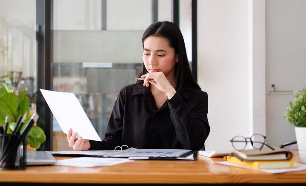 Młoda azjatycka kobieta biznesu siedzi w miejscu pracy i czyta dokument finansowy. - Zdjęcie, obraz