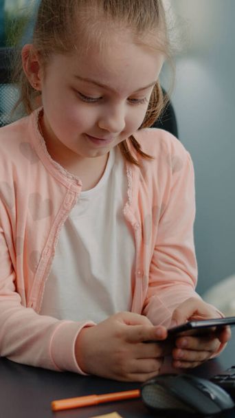 Маленька дівчинка дивиться на дисплей смартфона і посміхається за столом
 - Фото, зображення
