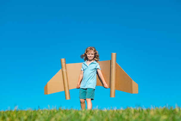 Happy child play with toy plane cardboard wings against blue sky. Kid having fun in summer field outdoor. Portrait of boy with paper wings. - Zdjęcie, obraz